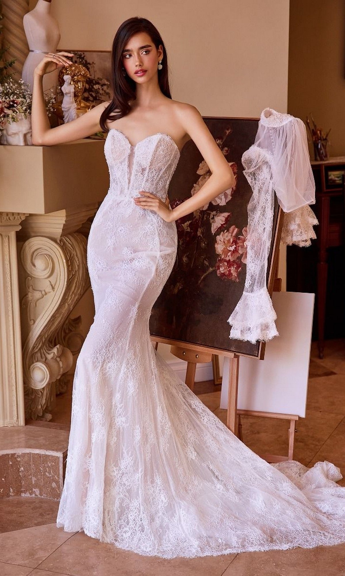 A woman wearing the Andrea and Leo WL042 long wedding dress with a fit-and-flare silhouette stands indoors beside a floral painting and an easel draped with a lace veil.